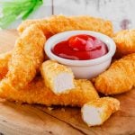 Chicken tenders on a brown cutting board with a small bowl of ketchup