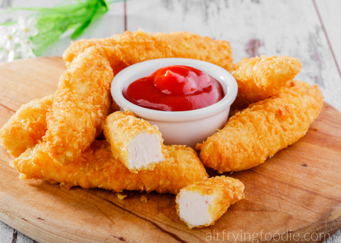 Chicken tenders on a brown board with a bowl of ketchup.