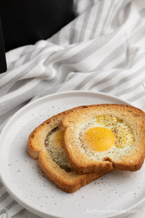 Eggs in a basket on a white plate with salt and pepper sprinkled over the top. Black Air Fryer in the background.