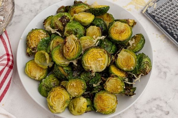 Brussel sprouts cut into halves, cooked in the air fryer, served on a white plate and topped with parmesan cheese. 