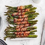 Golden brown crispy bacon wrapped around asparagus stalks on a white rectangle plate.