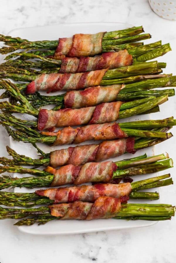 Golden brown crispy bacon wrapped around asparagus stalks on a white rectangle plate.