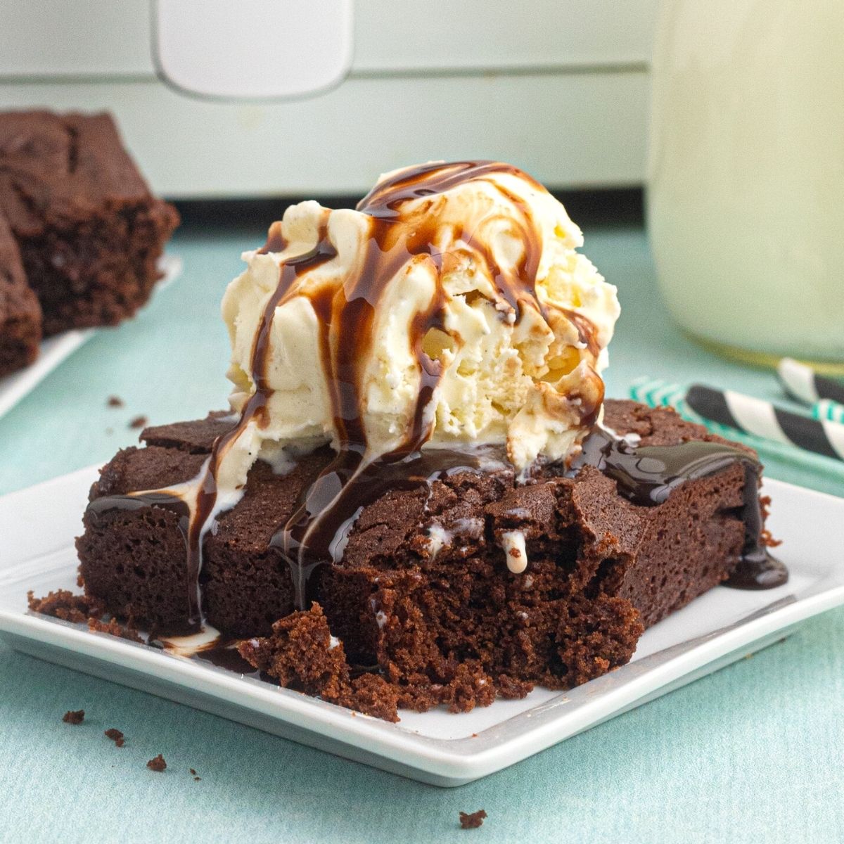 Air Fryer chocolate brownie on a white plate with ice cream and drizzled chocolate on top, with a bite out of brownie.