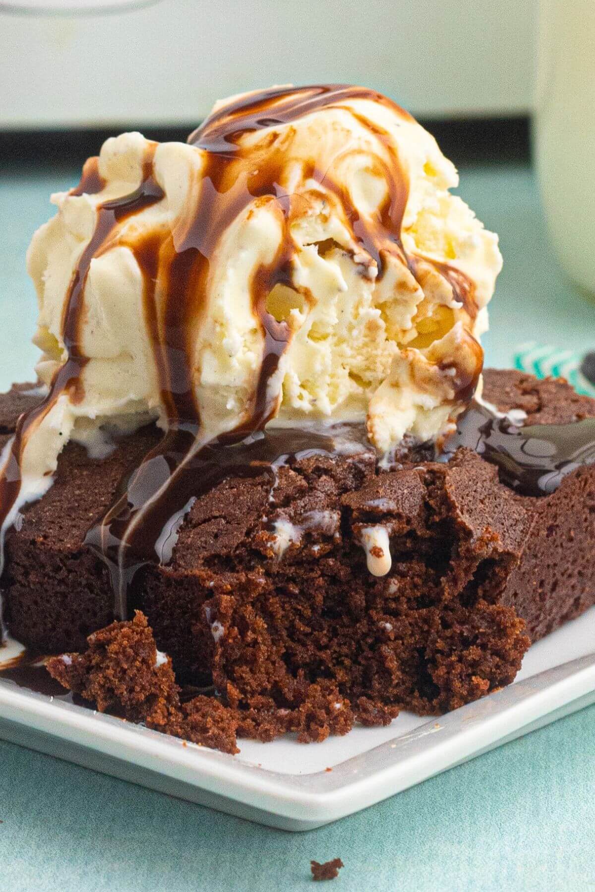 Chocolate brownie topped with vanilla ice cream and drizzled with chocolate syrup, on a white plate, in front of an air fryer, with a bite of the corner missing. 