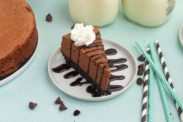 Slice of chocolate cheese cake on a white plate drizzled with chocolate and topped with whipped cream. 