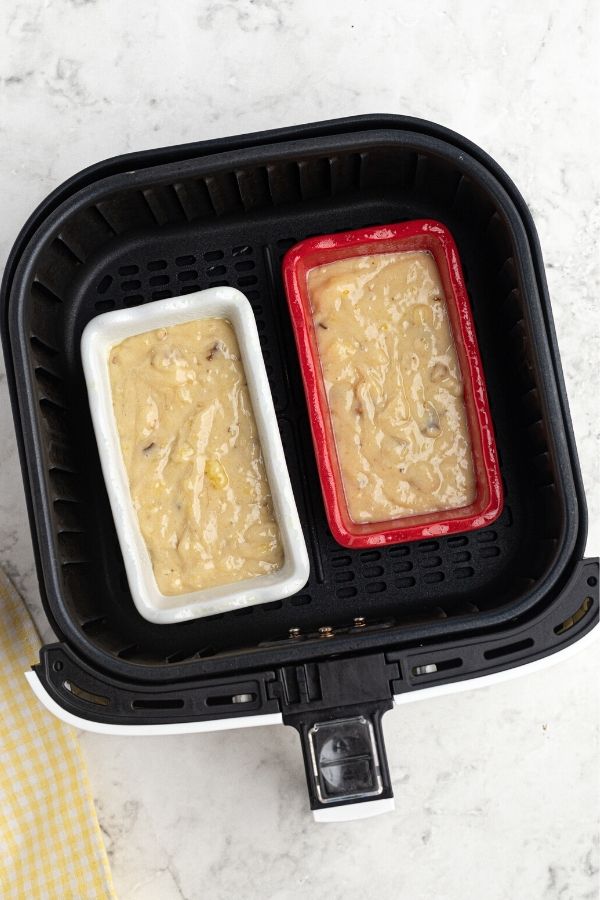 Uncooked loaves of bread in small loaf pans inside of an air fryer basket. One is red and the other is white. 