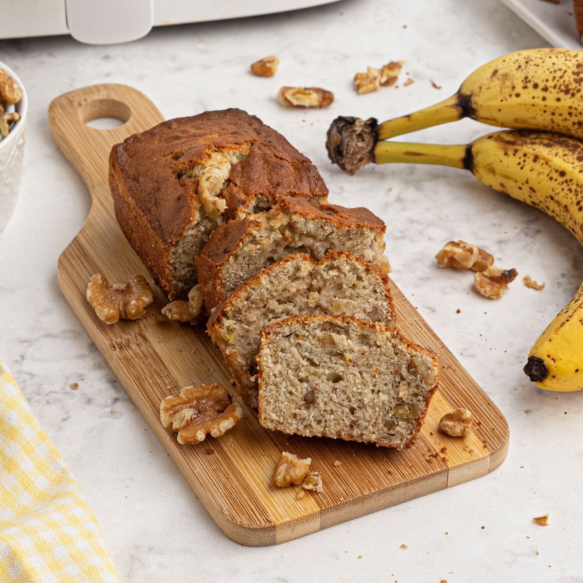 sliced loaf of banana nut bread in front of an air fryer next to bananas and scattered walnuts