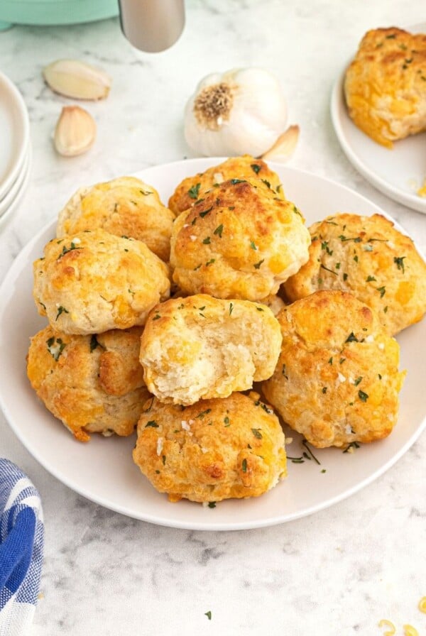 Golden cooked cheddar biscuits with parsley flakes on top, served on a white plate with shredded cheddar cheese and garlic cloves, scattered on the table.