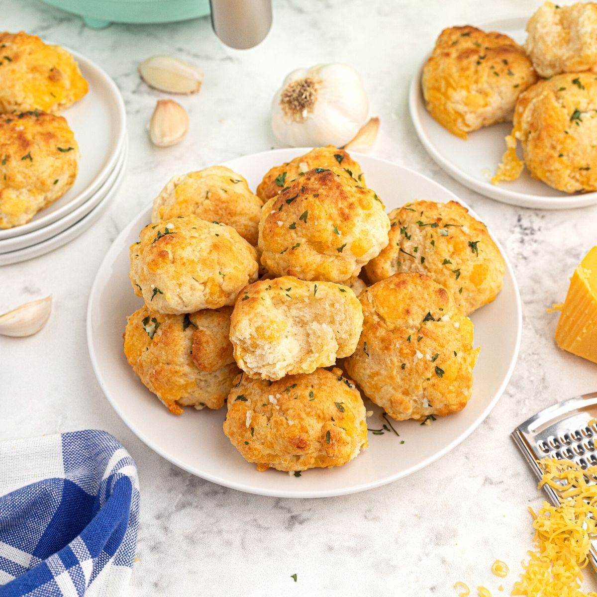 Golden cooked cheddar biscuits with parsley flakes on top, served on a white plate with shredded cheddar cheese and garlic cloves, scattered on the table.
