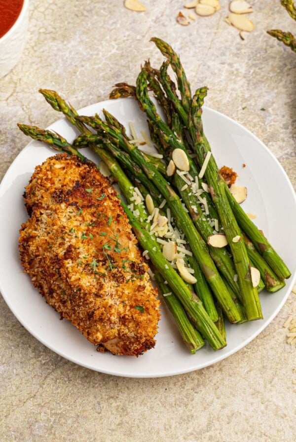 Cooked parmesan crusted chicken served with green asparagus on a white plate.