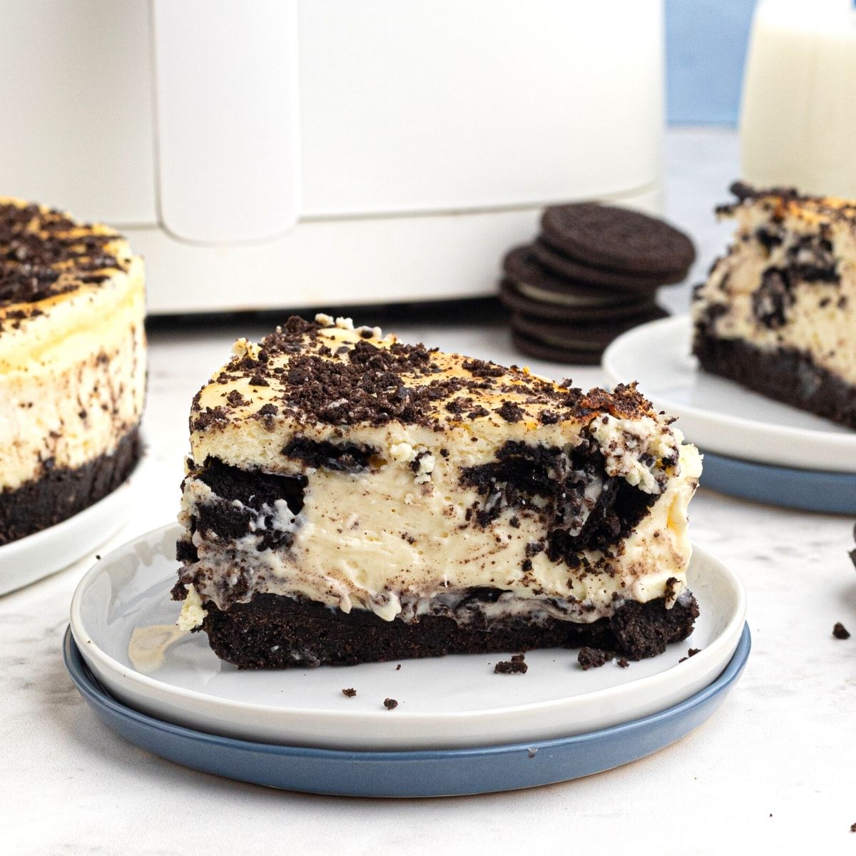 creamy slice of Oreo cheesecake served on a white plate in front of an air fryer.