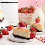 Creamy sliced piece of cheesecake, with a chocolate crust, served with strawberries on a white plate in front of an air fryer.
