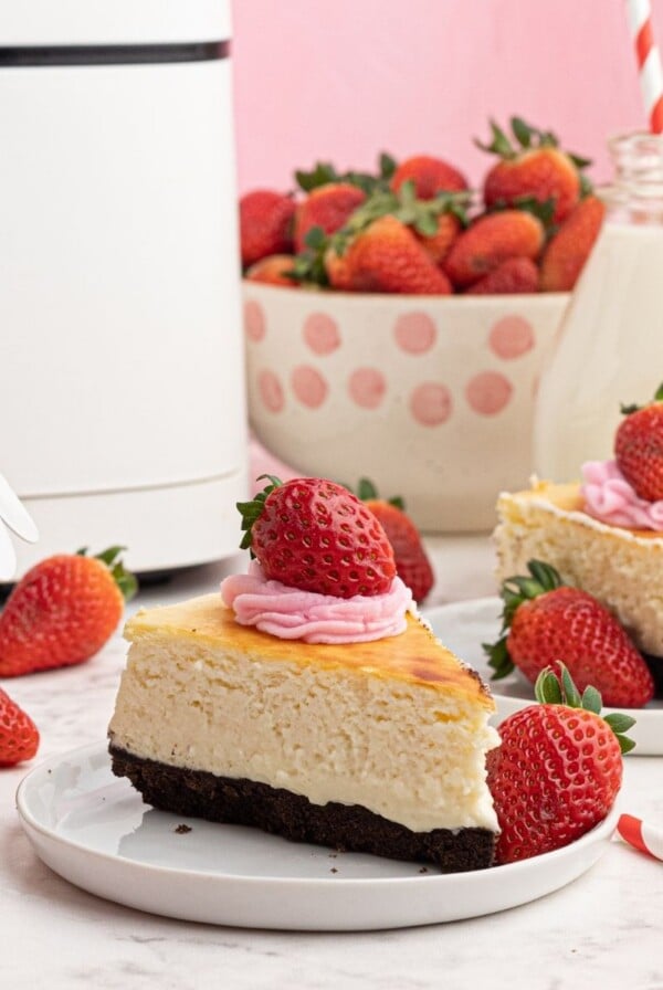 Creamy sliced piece of cheesecake, with a chocolate crust, served with strawberries on a white plate in front of an air fryer.