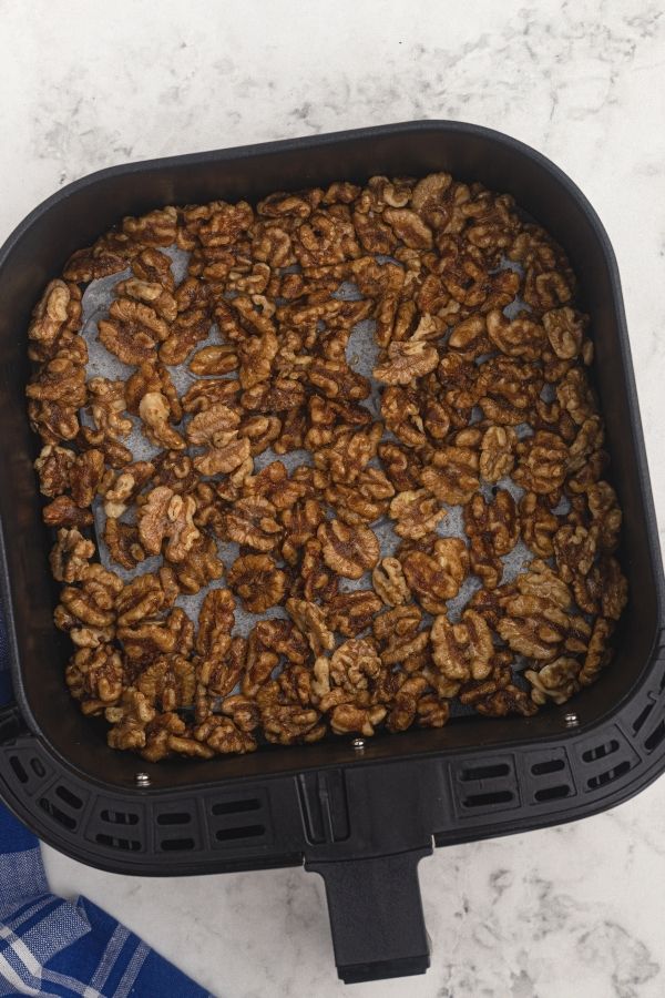 Uncooked walnuts in an air fryer basket, seasoned with melted butter, sugars, and the other ingredients. 
