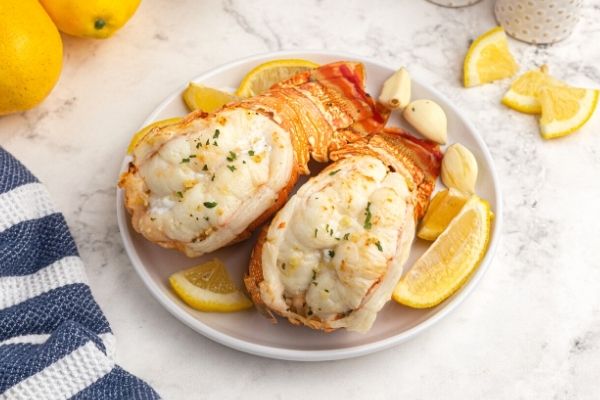 Cooked juicy lobster tails, served on a white plate with lemon wedges on the table. 
