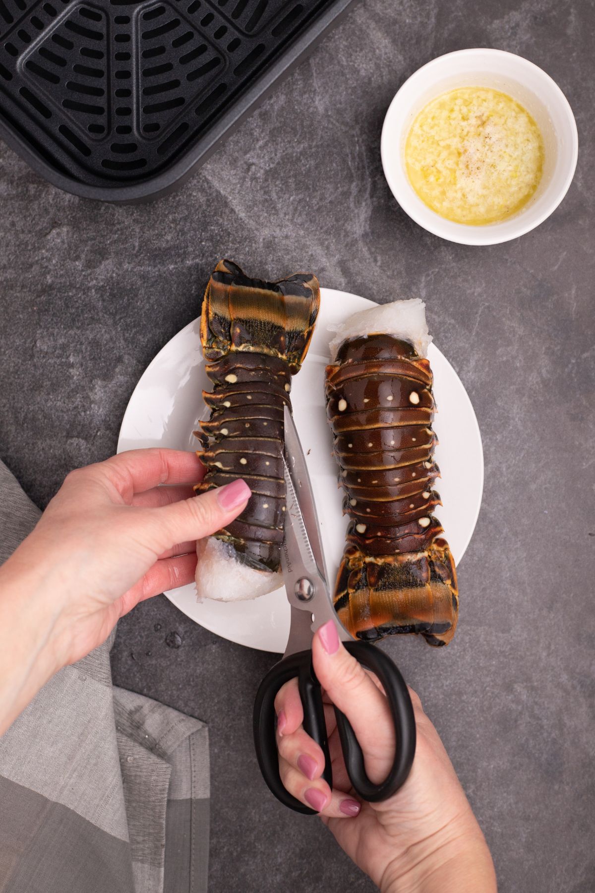 Lobster tails being cut open with kitchen shears on a white plate.