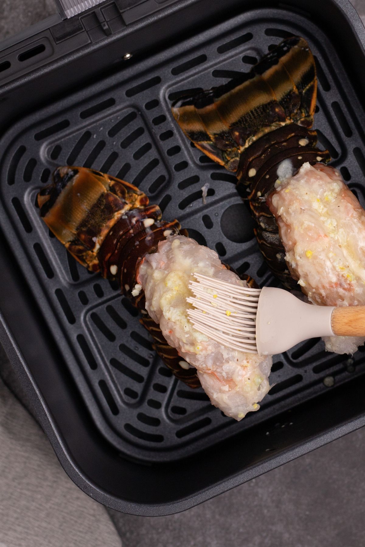 Lobster tails in an air fryer basket being brushed with melted butter and seasonings. in air fryer basket.