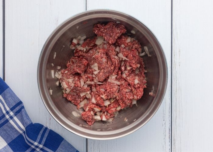 Raw  hamburger meat combined with chopped onions and seasonings in a bowl on a white wooden table.  