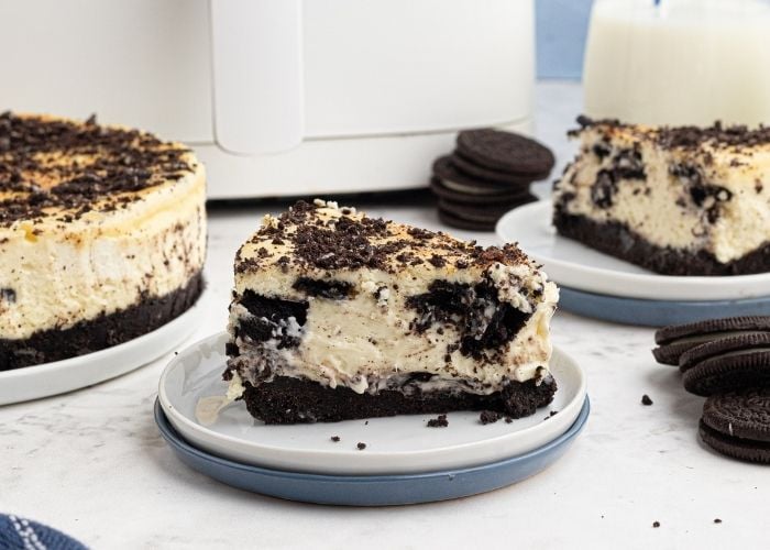 Air Fryer Oreo Cheesecake, cut and sliced on a plate in front of an air fryer with Oreo's spread around it. 
