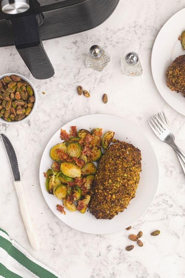 Pistachio crusted chicken cut into slices, served on a white plate next to Brussel sprouts. In front of an air fryer, with pistachios scattered on the table. 
