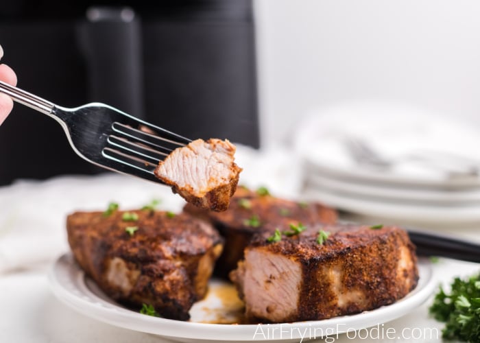 Bite of pork chop made in the air fryer on a fork, with pork chops on a plate and air fryer in the background. 
