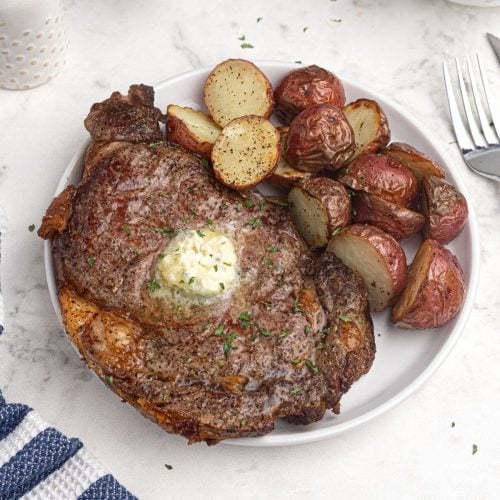 Juicy and cooked ribeye steak on a while plate. Served with red potatoes.