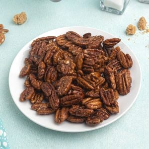 Cooked pecans, served on a white plate, in front of an air fryer, with scattered pecans and brown sugar around the plate.