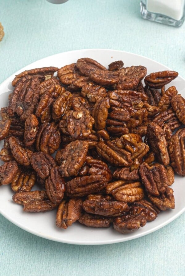 Cooked pecans, served on a white plate, in front of an air fryer, with scattered pecans and brown sugar around the plate.