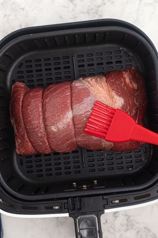 Uncooked top round roast in the air fryer basket, being brushed with olive oil, and seasoned with salt and pepper. 