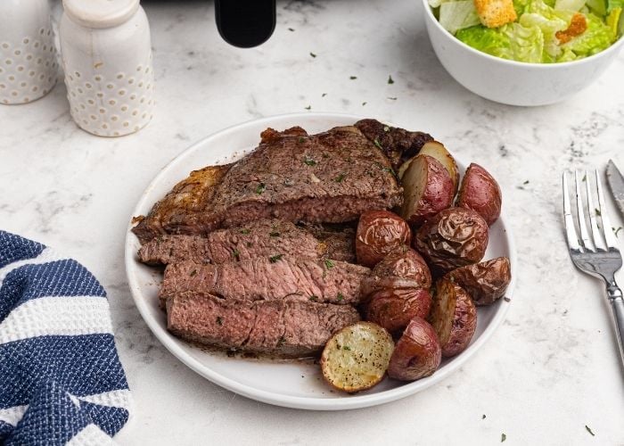 Juicy, cooked and sliced ribeye steak, in front of an air fryer, served on a white plate, with potatoes. 