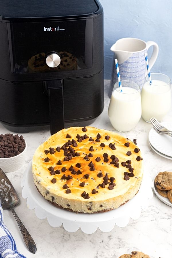 Whole creamy cooked chocolate chip cheesecake served on a white stand. Sitting in front of the air fryer. 