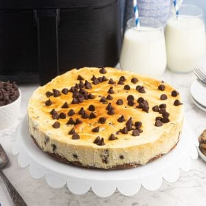 Air Fryer Chocolate Chip cheesecake on a white cake stand, in front of an air fryer.