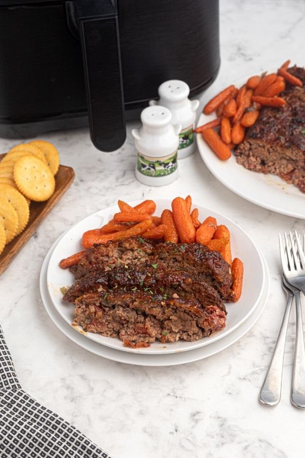 Cooked and sliced meatloaf on a white plate with baby carrots, in front of an air fryer. 