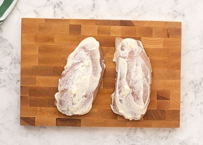 Uncooked chicken on a cutting board, seasoned with salt and pepper, then covered in mayonnaise. 