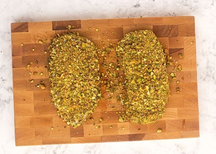 Uncooked chicken on a cutting board, seasoned with salt and pepper, then covered in mayonnaise and chopped pistachios.