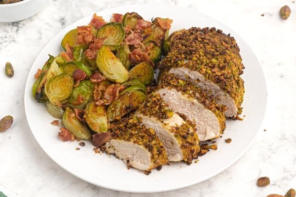 Pistachio crusted chicken cut into slices, served on a white plate next to Brussel sprouts. 
