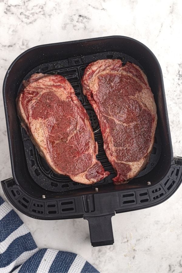 Juicy and thick uncooked ribeye steaks in an air fryer basket, coated with olive oil and seasonings. 