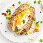Baked potato stuffed with cheese, sour cream, bacon crumbles, on a white plate with chopped green onion and crumbles scattered on the table.