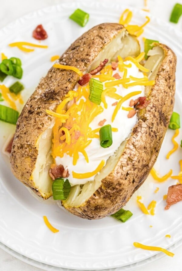 Baked potato stuffed with cheese, sour cream, bacon crumbles, on a white plate with chopped green onion and crumbles scattered on the table.