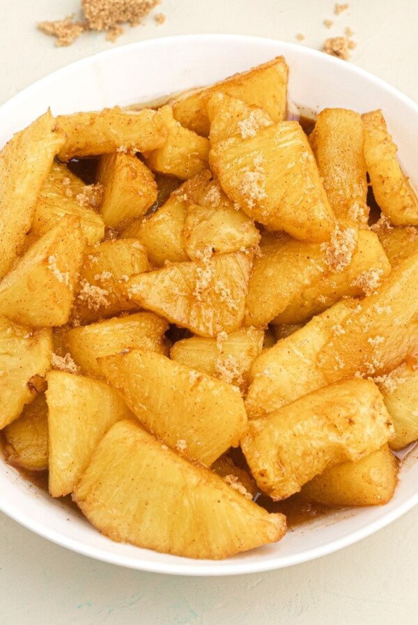 Golden juicy pineapple bites, in juices, served in a white bowl, topped with sprinkled brown sugar.