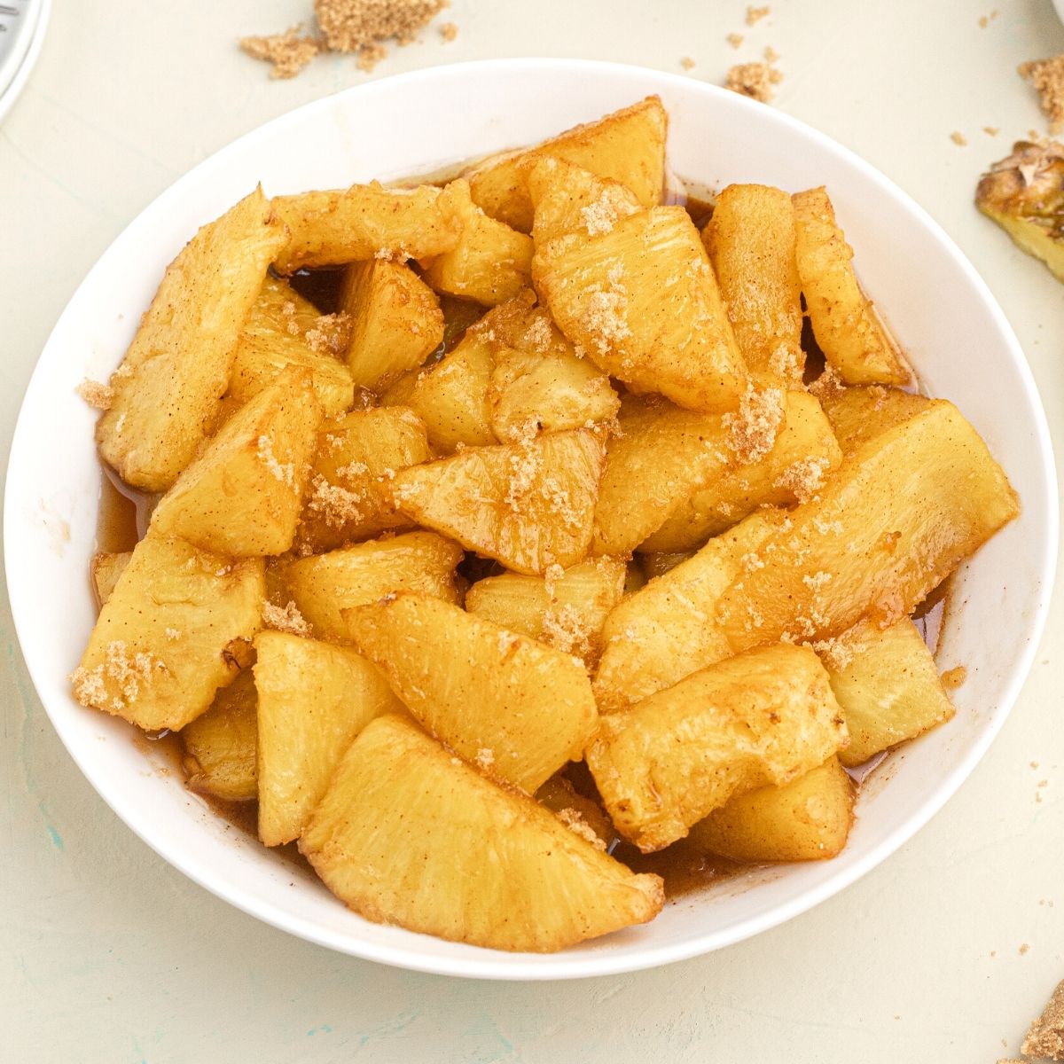 Golden juicy pineapple bites, in juices, served in a white bowl, topped with sprinkled brown sugar.