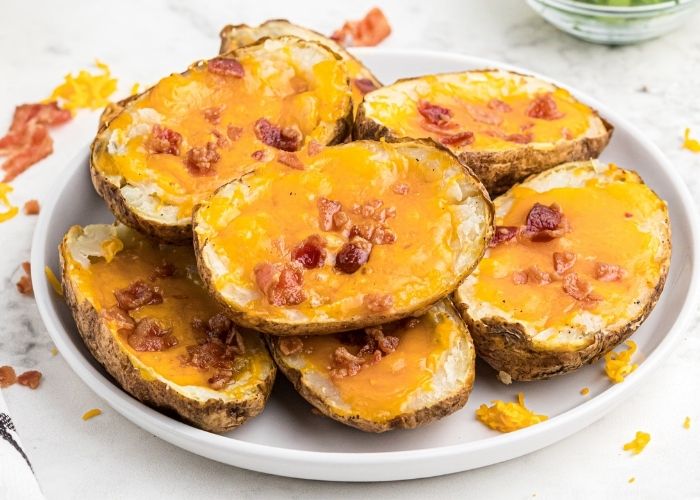 Cooked potato skins with melted cheddar cheese and bacon crumbles on top, served on a white plate with crumbled bacon. 