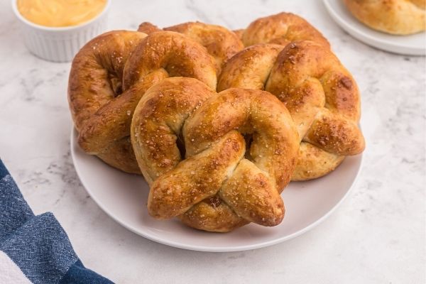 Golden soft pretzels served on a white plate. topped with salt and a small bowl of melted cheese dipping sauce. 