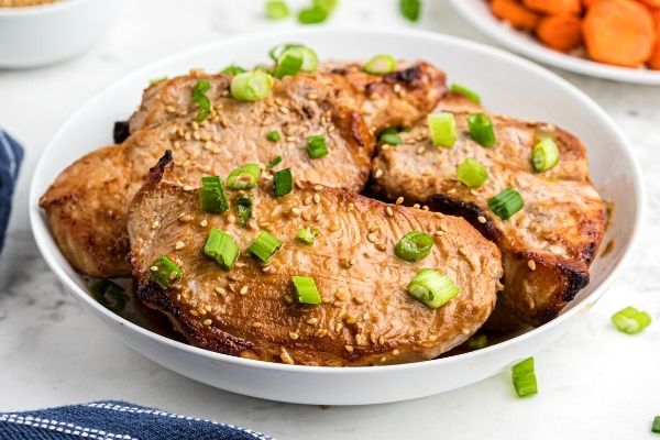 Juicy teriyaki pork chops served in a white bowl, with chopped green onions and sprinkles of sesame seeds on top. 
