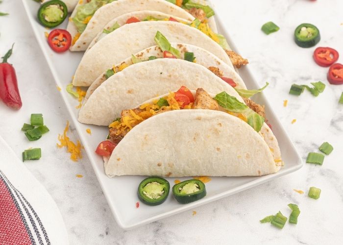 flour tortillas, filled with seasoned chicken, served on a white rectangle plate. Chopped vegetables and topped with lettuce, tomatoes, shredded cheese, and jalapenos. 