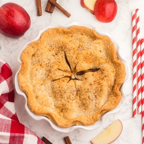 Golden baked apple pie, with red apples and red and white straws on the table.