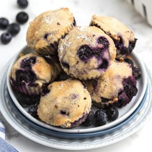 Blueberry muffins stacked on a white plate with fresh blueberries scattered around the table.