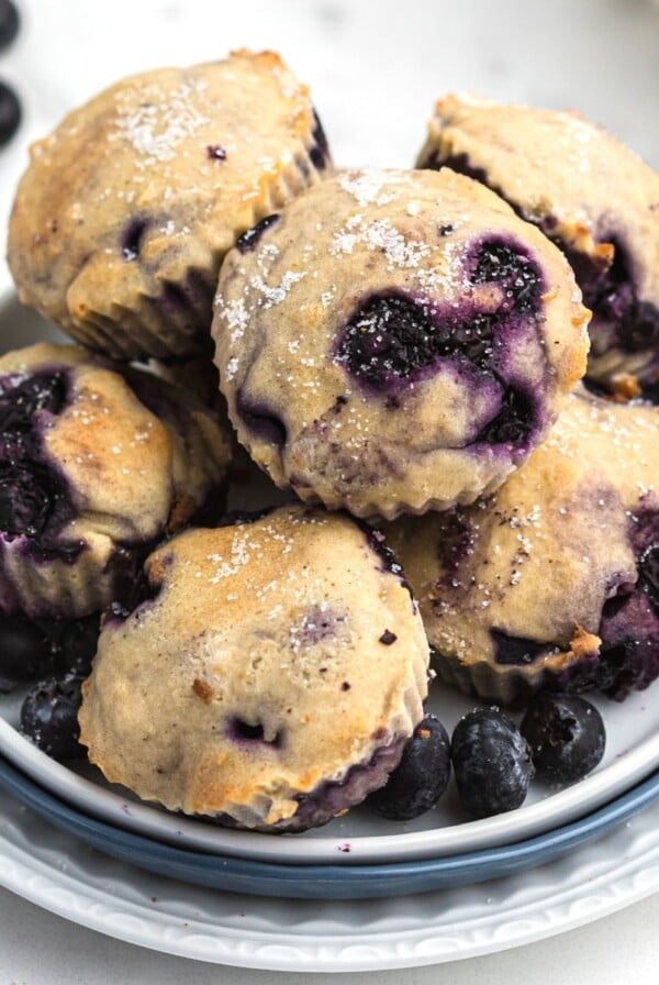 Blueberry muffins stacked on a white plate with fresh blueberries scattered around the table.