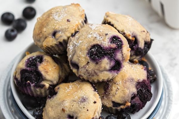 Moist blueberry muffin bites stacked on a white plate, sprinkled with sugar and fresh blueberries on the table behind it. 