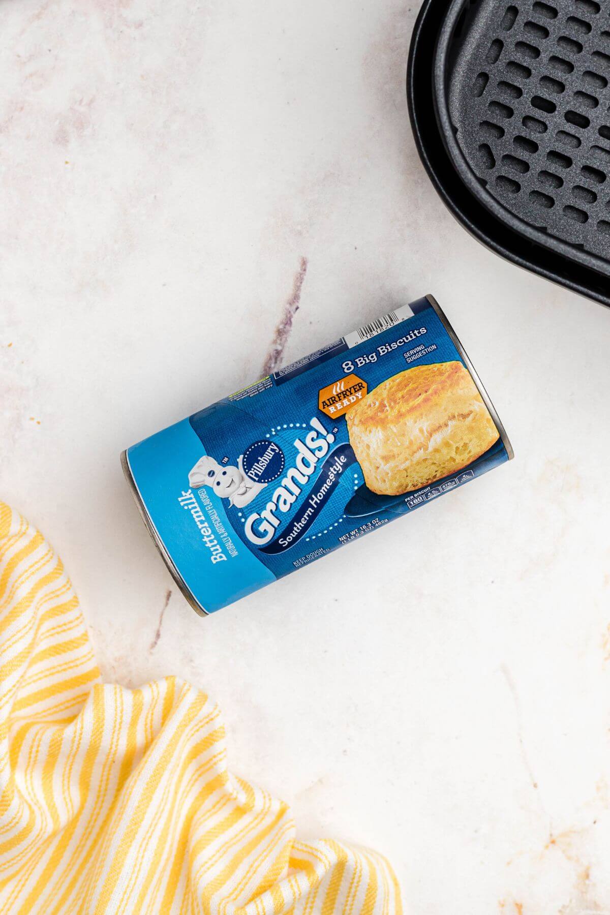 Can of grands biscuits on marble table in front of the air fryer basket. 